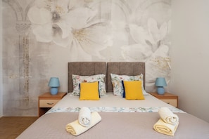 Quarto com cama, duas mesinhas de cabeceira, um mural floral na parede e duas toalhas enroladas.