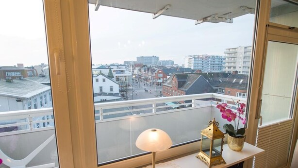 Quarto com vista para a paisagem urbana através de grandes janelas, uma pequena mesa com abajur, um objeto decorativo e um vaso de planta.
