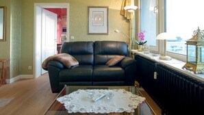 Sala de estar com sofá de couro preto, toalha de mesa de renda e vista para a cidade através de uma janela.