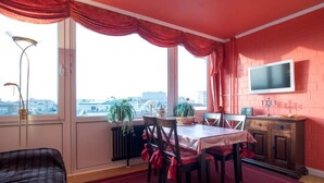 Quarto com mesa de jantar, cadeiras, televisão na parede e janela com cortinas.