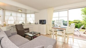 Een ruime woonkamer met een grijze bank, een salontafel en een televisie. Er is een eethoek met een tafel en stoelen, en uitzicht op de tuin door grote ramen.