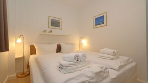 Een net opgemaakt bed met witte lakens en opgerolde handdoeken, twee bedlampen en twee ingelijste foto's aan de muur.