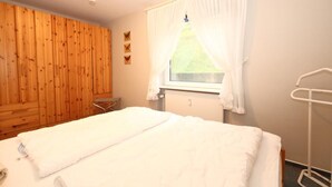 Une chambre avec une armoire en bois, un lit avec une literie blanche, une fenêtre avec des rideaux blancs et une table de chevet.