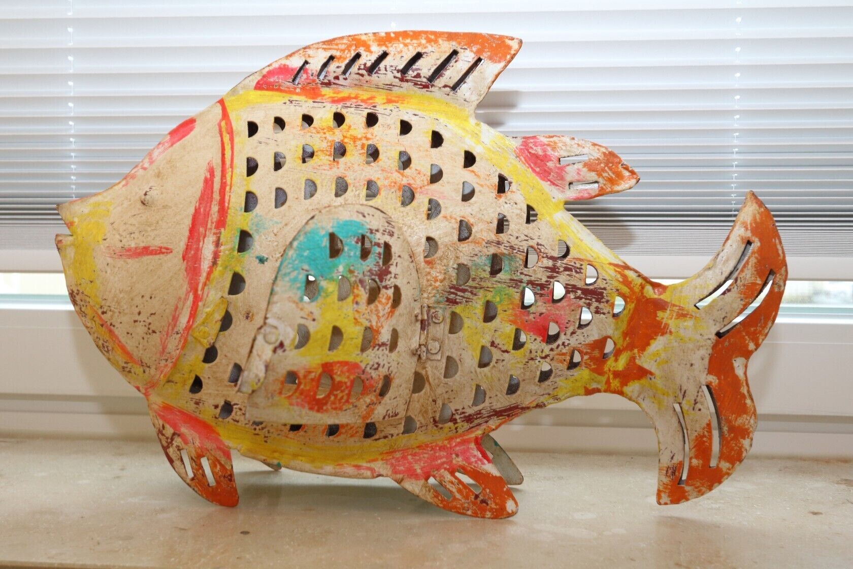 Une sculpture colorée représentant un poisson, percée de trous et dotée d’un bec, placée sur un rebord de fenêtre.
