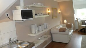 Una cocina compacta con microondas, tostadora y cafetera sobre estantes, un lavamanos y un sofá blanco en una sala de estar con lámpara y sillón.