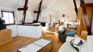 Un vaste espace de vie comprenant un canapé, une table basse et une table à manger. Il y a également un coin cuisine avec un évier et une cuisinière. La pièce est dotée de poutres en bois au plafond et d’une fenêtre avec des stores.