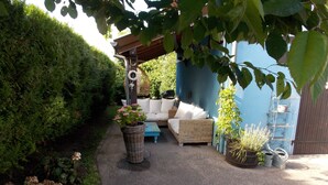 Un espace extérieur aménagé, abrité et doté d'un canapé en osier et de coussins, entouré de verdure et de plantes en pot.