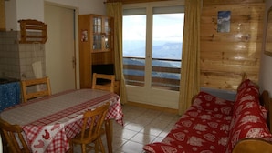 A cozy room with a dining table, chairs, a sofa, a TV, and a sliding glass door leading to a balcony with a mountain view. - 50m from ski slopes, balcony, parking, tv, ski locker, 30-32m², Les 7 Laux (Les Adrets)