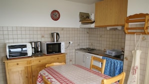 A small kitchen with a microwave, toaster, kettle, and stove. There is a wooden cabinet, a table with a checkered tablecloth, and a chair. - 50m from ski slopes, balcony, parking, tv, ski locker, 30-32m², Les 7 Laux (Les Adrets)