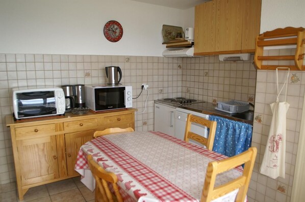 Une petite cuisine équipée d’un four à micro-ondes, d’un grille-pain, d’une bouilloire et d’une cuisinière. On y trouve un meuble en bois, une table recouverte d’une nappe à carreaux et une chaise.