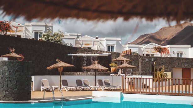 A pool area with lounge chairs and umbrellas, a stone wall, and white buildings with a mountain backdrop. - Apartamentos Livvo Coloradamar (Yaiza)