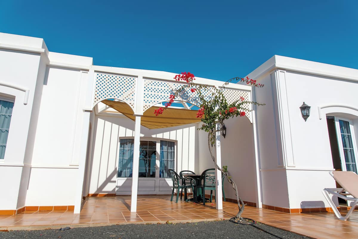 Un bâtiment blanc doté d’un patio abrité, d’une petite table avec des chaises et d’un pot de fleurs orné de fleurs rouges.
