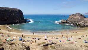 A beach with rocky cliffs, clear blue water, and people sunbathing. - Apartamentos Livvo Coloradamar (Yaiza)