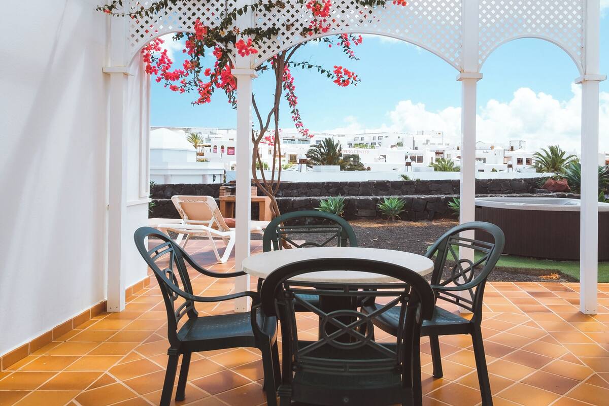 Une terrasse avec une table et des chaises, une pergola ornée de fleurs et, en arrière-plan, une vue sur des bâtiments.