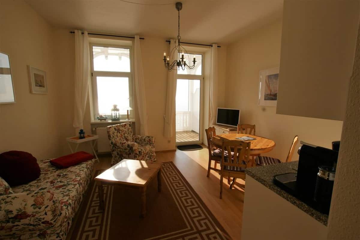 A cozy living room with a sofa, coffee table, and a dining area with chairs and a table. There is a TV and a small table with a lamp.