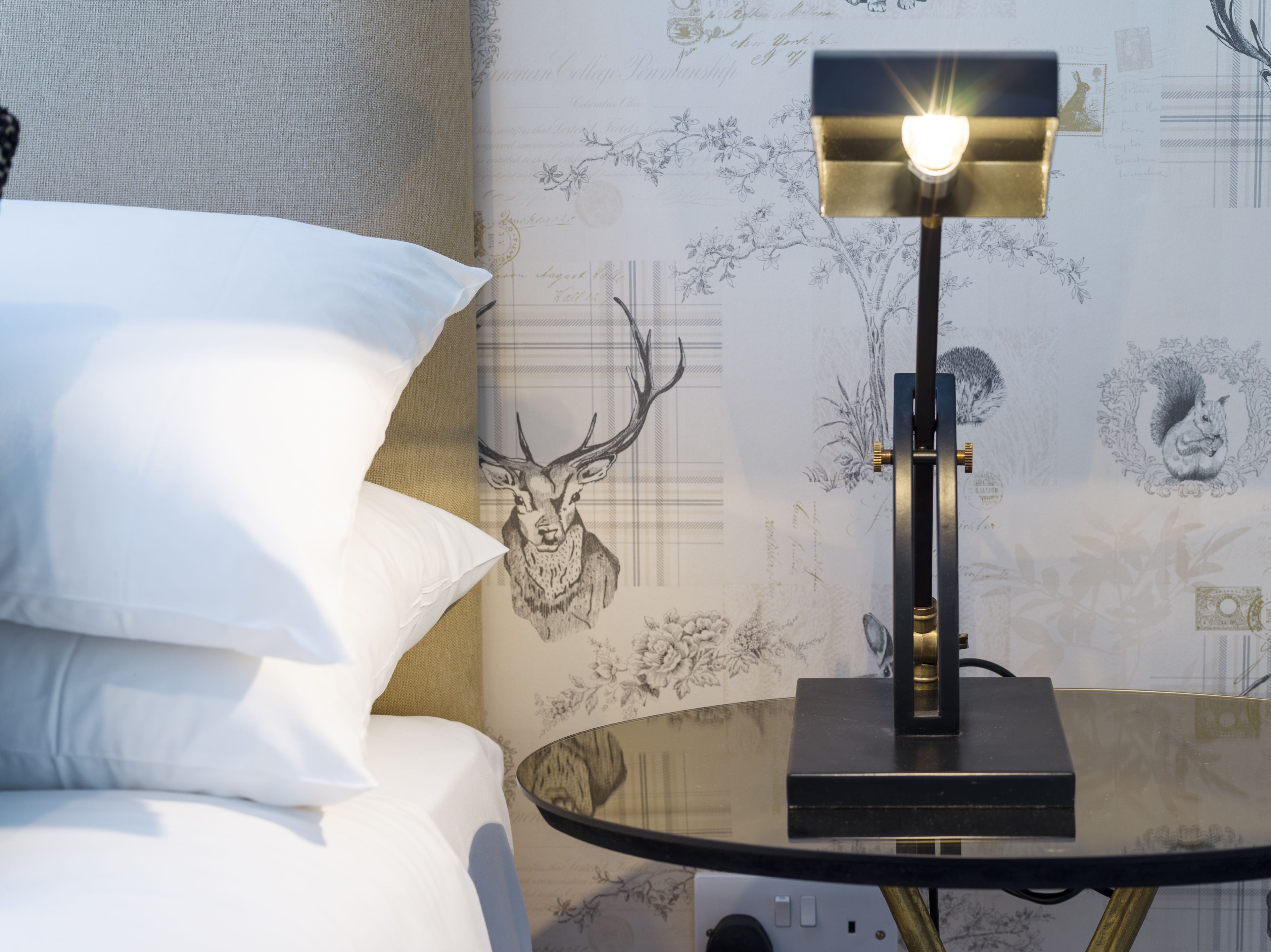 Una cama con almohadas blancas, una mesa de vidrio con una lámpara y una pared con una ilustración de un ciervo.