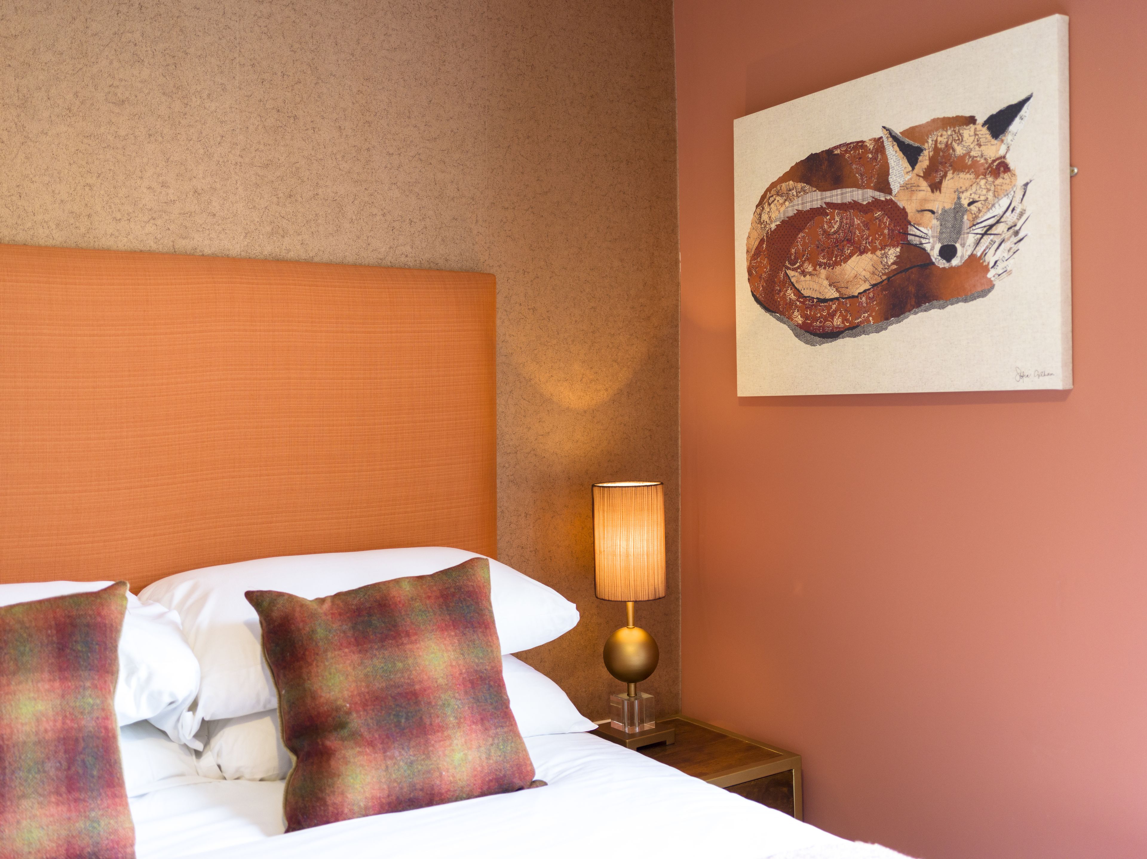 Una cama bien hecha con sábanas blancas y almohadas de colores, una lámpara de pared y un cuadro de un zorro en la pared.
