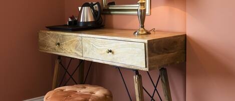 A wooden desk with a lamp, a mirror, and a tufted ottoman in a room with a pink wall.