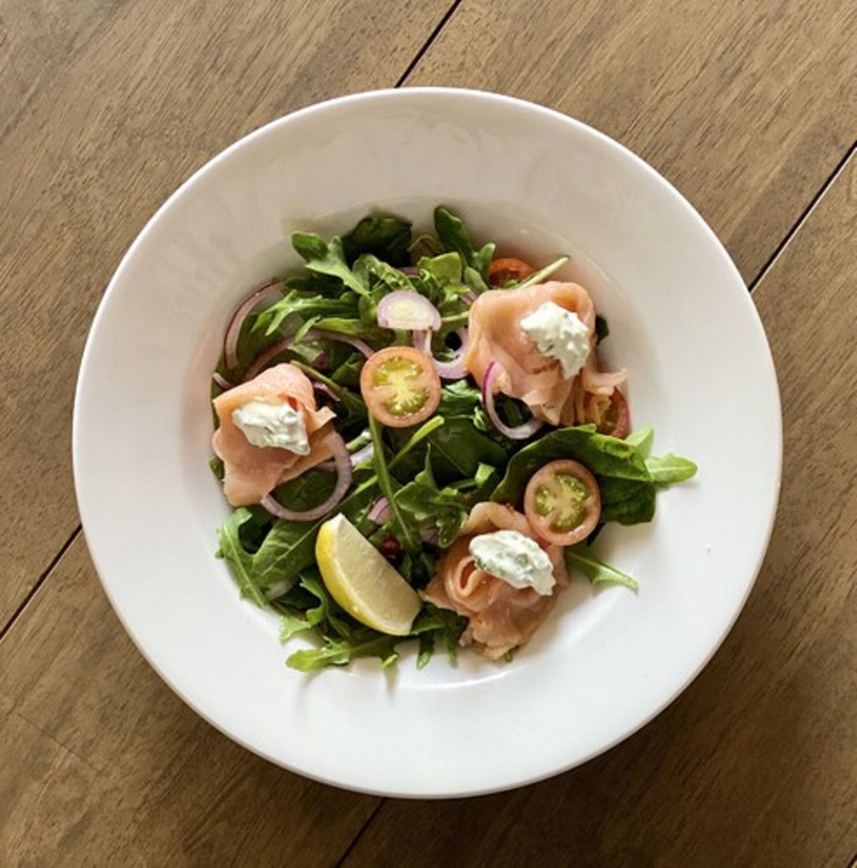 A white bowl of salad with spinach, sliced tomatoes, red onions, and smoked salmon, garnished with a lemon wedge.