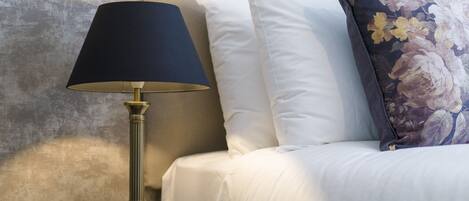 A bed with white bedding, a dark-colored lamp, and a floral pillow.