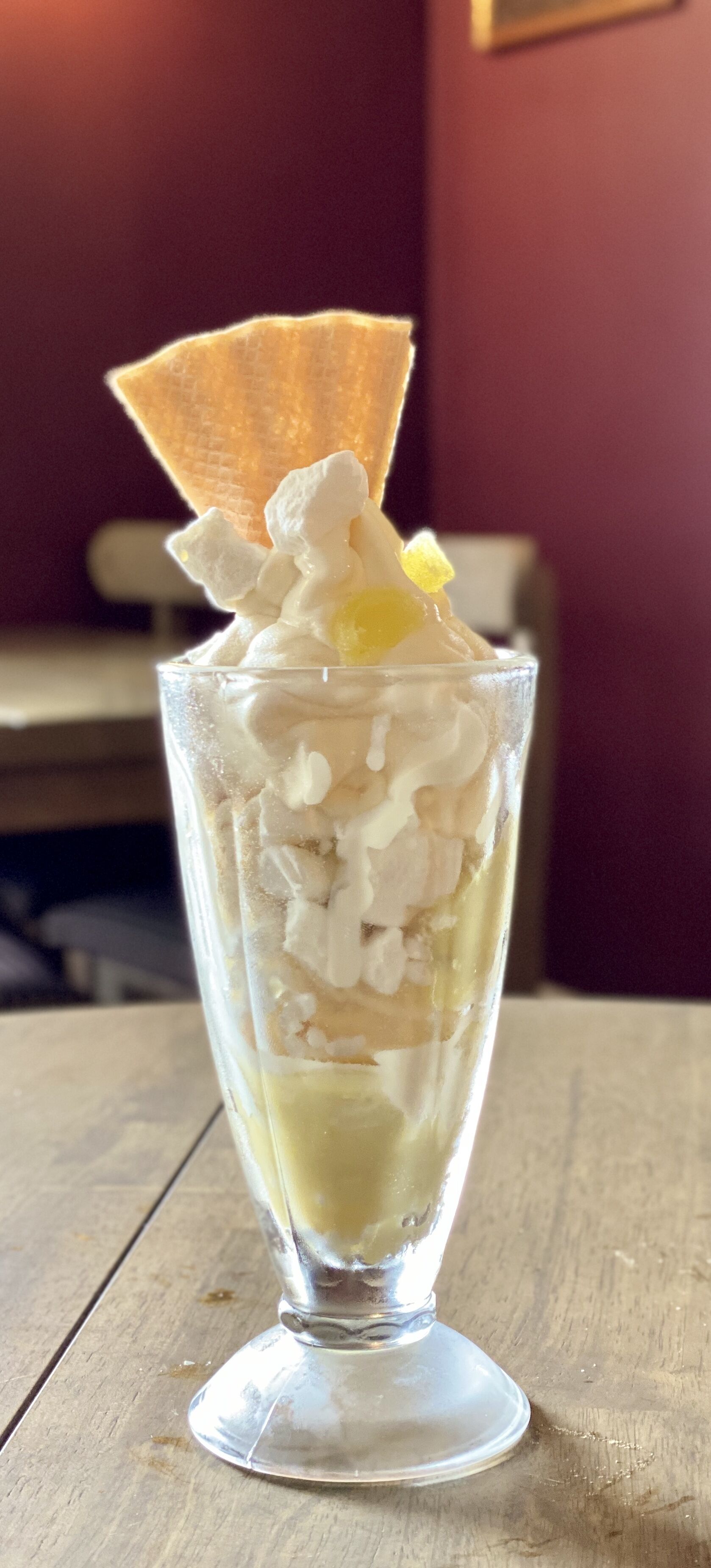 Un postre en un vaso alto con crema batida y una galleta encima, sobre una mesa de madera.