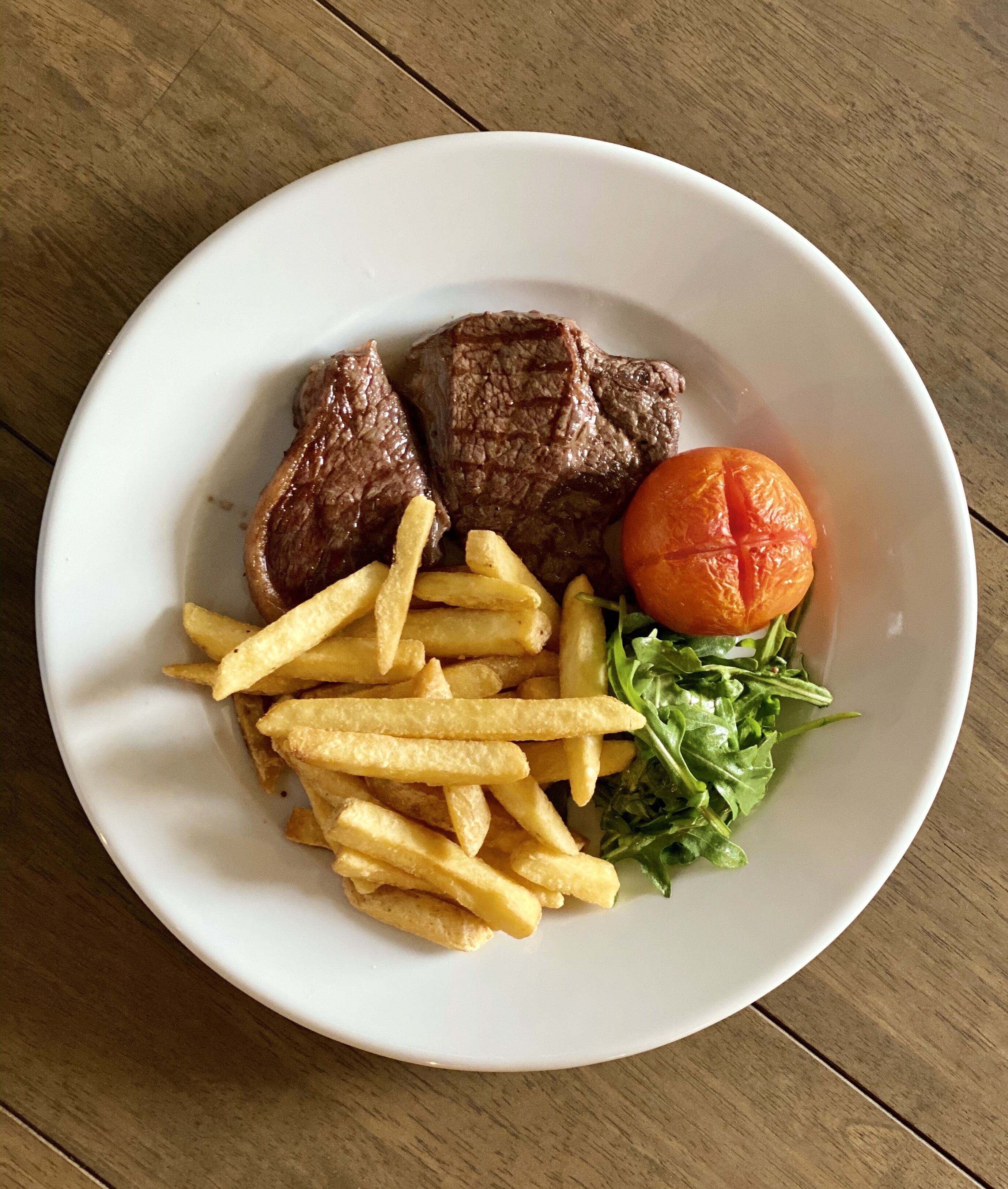 Une assiette blanche contenant deux morceaux de viande, des frites, une tomate et quelques légumes verts, le tout posé sur une table en bois.