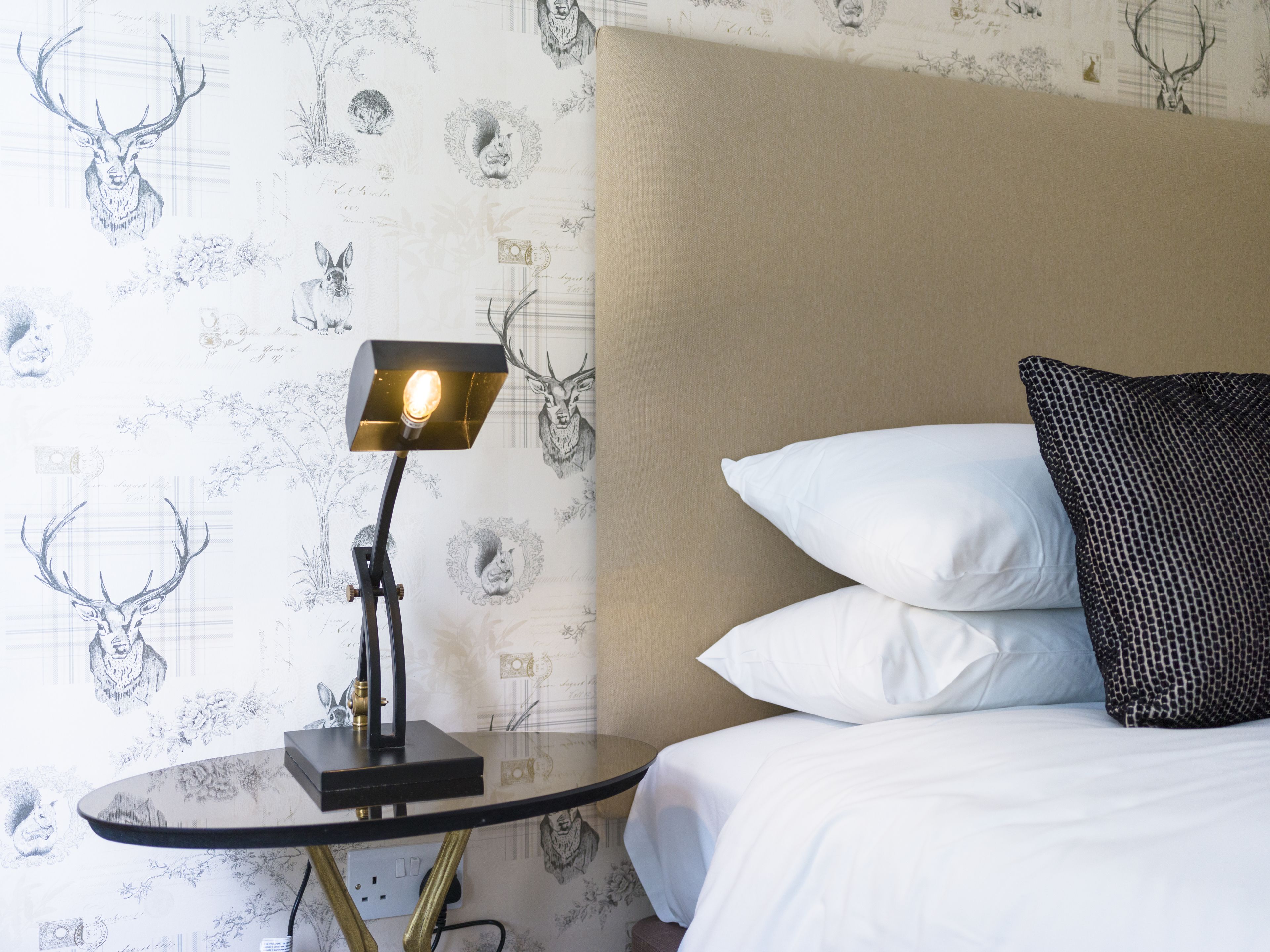 A neatly made bed with white and patterned pillows, a bedside lamp, and a wall with a deer and floral wallpaper.