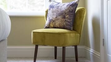 Een gele fauteuil met een bloemenkussen in een kamer met raam en vloerbedekking.