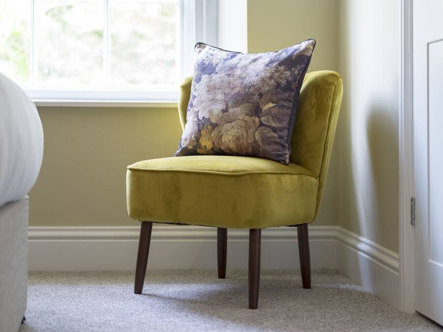 Een gele fauteuil met een bloemenkussen in een kamer met raam en vloerbedekking.