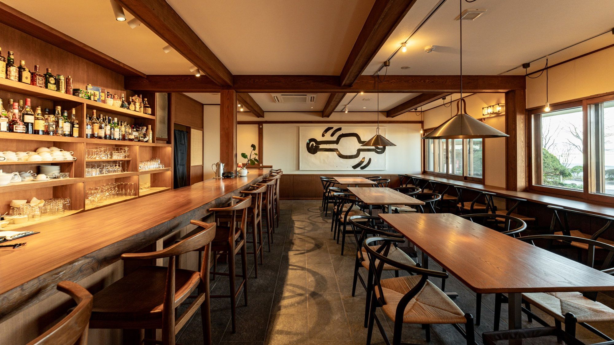 Zona de bar con estanterías de madera llenas de botellas, vasos y platos. Varias mesas y sillas dispuestas para sentarse.