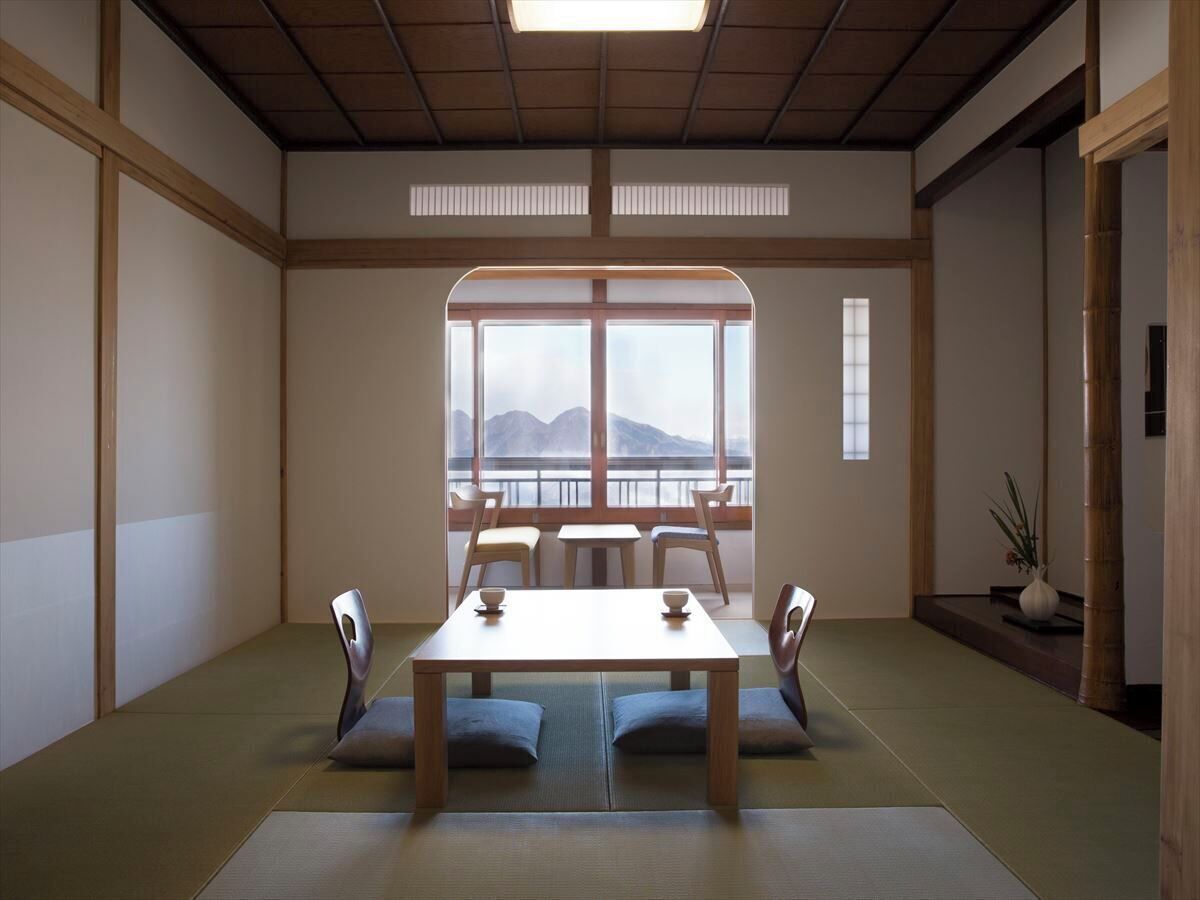 A traditional Japanese room with tatami flooring, a low table with cushions, and a view of mountains through large windows.