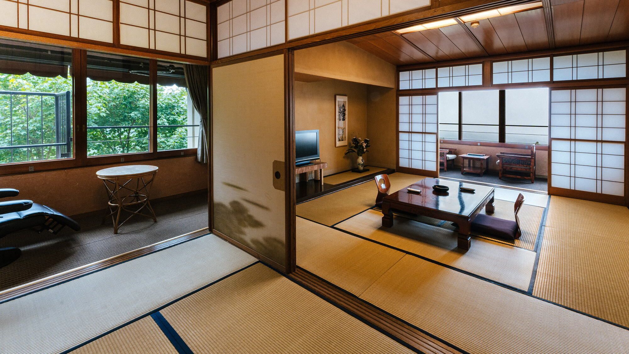 A traditional Japanese room with tatami flooring, a tatami mat table, a low wooden table, a television, and a view of a garden through large windows.