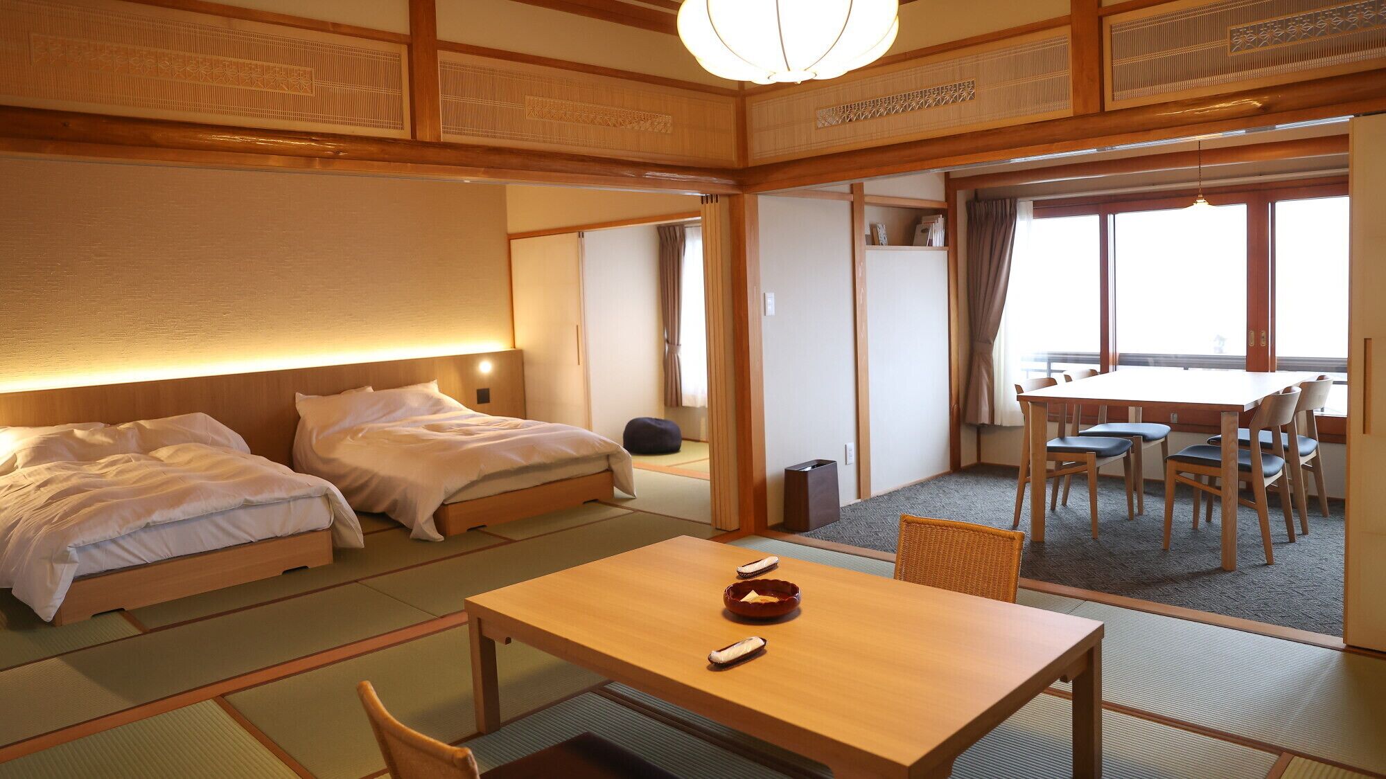 A traditional Japanese-style hotel room with tatami flooring, a wooden bed, a table with a remote control, and a dining area with chairs.