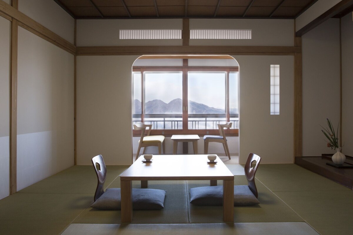 A traditional Japanese room with tatami flooring, a low table, and a view of mountains through large windows.