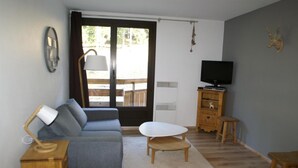 A living room with a sofa, a small table, a TV on a wooden cabinet, and a sliding glass door leading to an outdoor area. - 50m from ski slopes, balcony, parking, tv, ski locker, 28m², Les 7 Laux (Les Adrets)