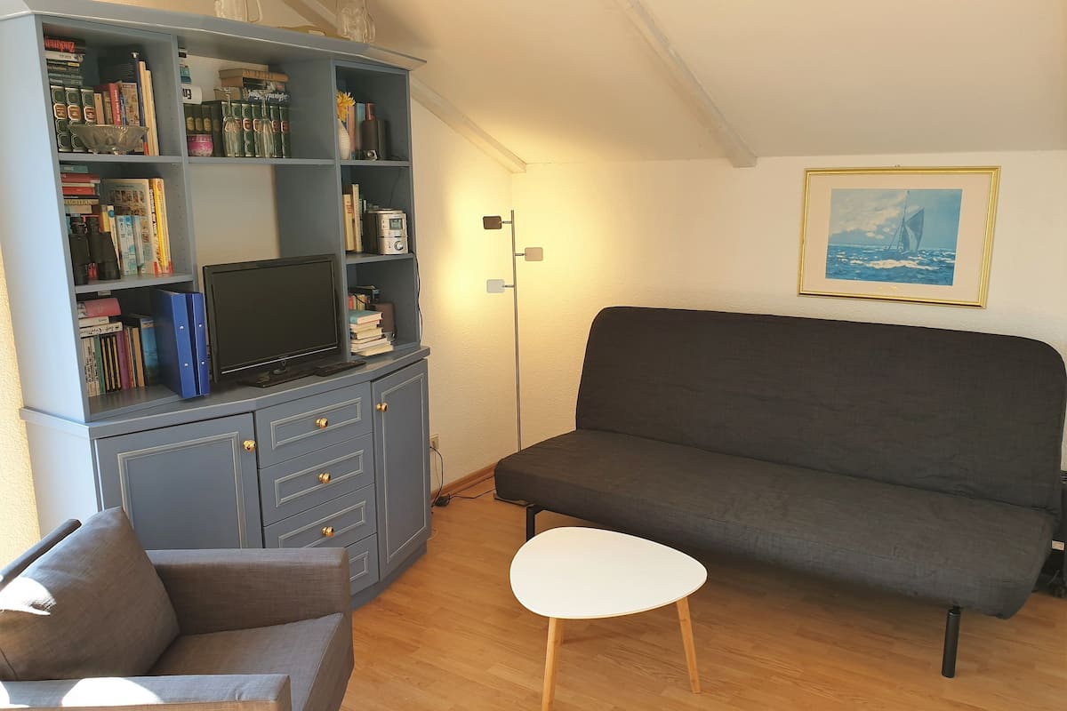 Un salón con un sofá gris, una mesita blanca, un aparador gris con libros y un televisor, y un cuadro en la pared.
