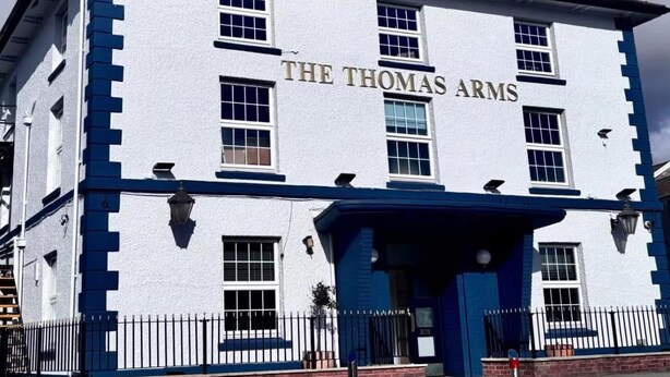 Un edificio con el nombre "The Thomas Arms" en su fachada, que presenta un estilo arquitectónico tradicional, una entrada destacada y una combinación de colores azul y blanco.