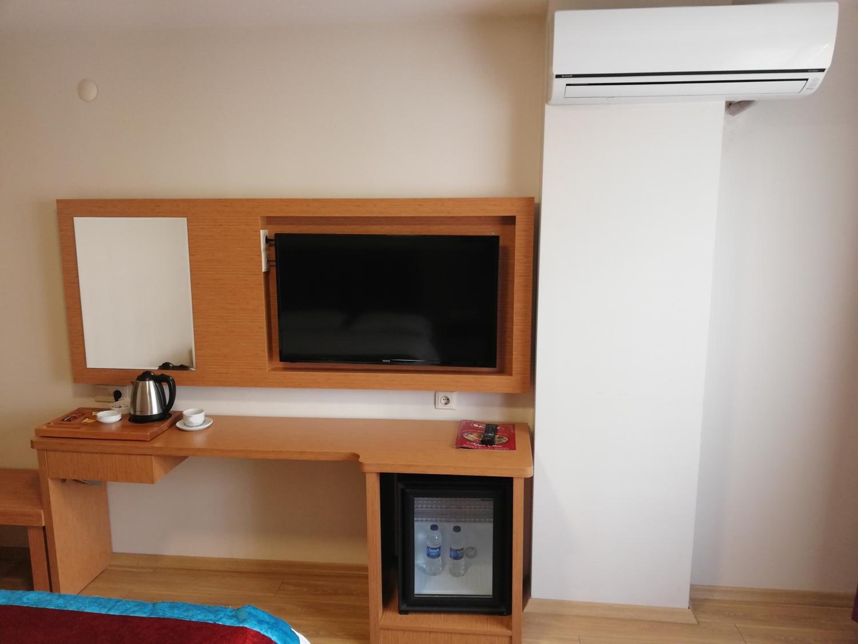 A hotel room with a wooden desk, a television, a mini-fridge, and an air conditioning unit.