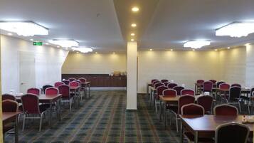 Un salón de conferencias con filas de mesas y sillas, suelo alfombrado e iluminación en el techo.