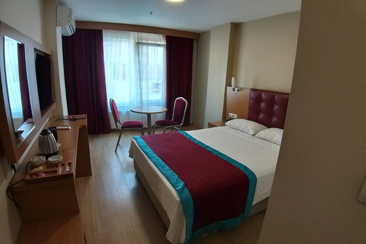 A hotel room with a bed, a desk with a kettle, a chair, a TV, and a window with curtains.