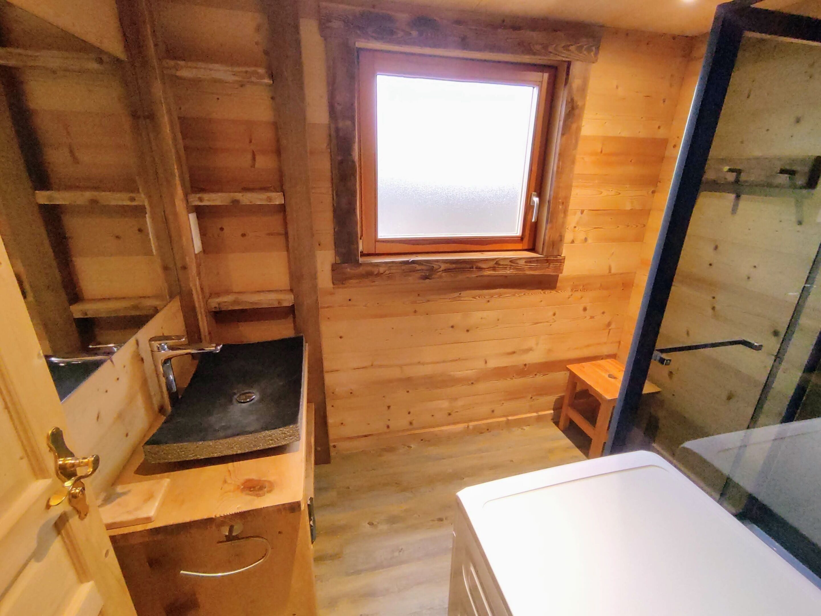 L’intérieur d’une cabane en bois, avec une fenêtre, un petit tabouret en bois et une porte en métal.