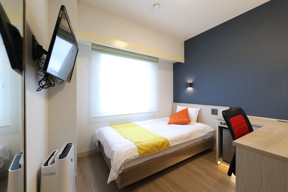 A hotel room with a bed, a desk, a chair, a television mounted on the wall, and a window.
