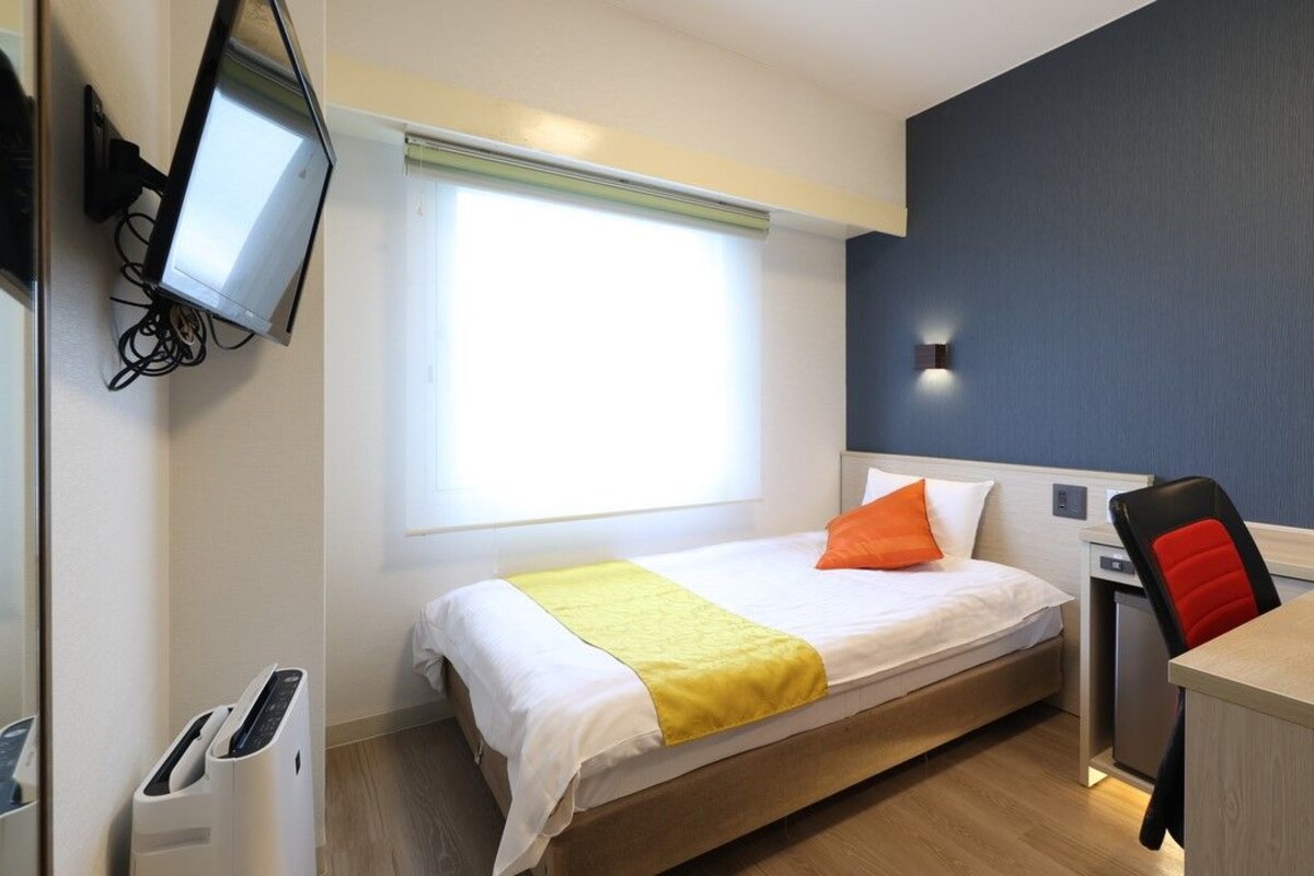 A hotel room with a bed, a desk, a chair, a TV mounted on the wall, and a window.