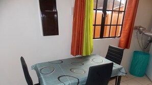 Ein Essbereich mit einem Tisch, der mit einer gemusterten Tischdecke bedeckt ist, zwei Stühle, einem Fenster mit orangefarbenen Vorhängen und einem gefliesten Boden.