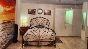 A bedroom with a bed, a nightstand, a wall-mounted lamp, a ceiling fan, a chandelier, a wardrobe, and a painting of a sunset. - Stewart Hotel Apt - Trincity, Airport, Full Apt (Trincity)