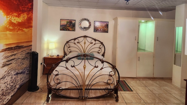 Um quarto com uma cama, uma mesa de cabeceira, uma luminária na parede, um ventilador de teto, um lustre, um guarda-roupa e uma pintura de um pôr do sol.