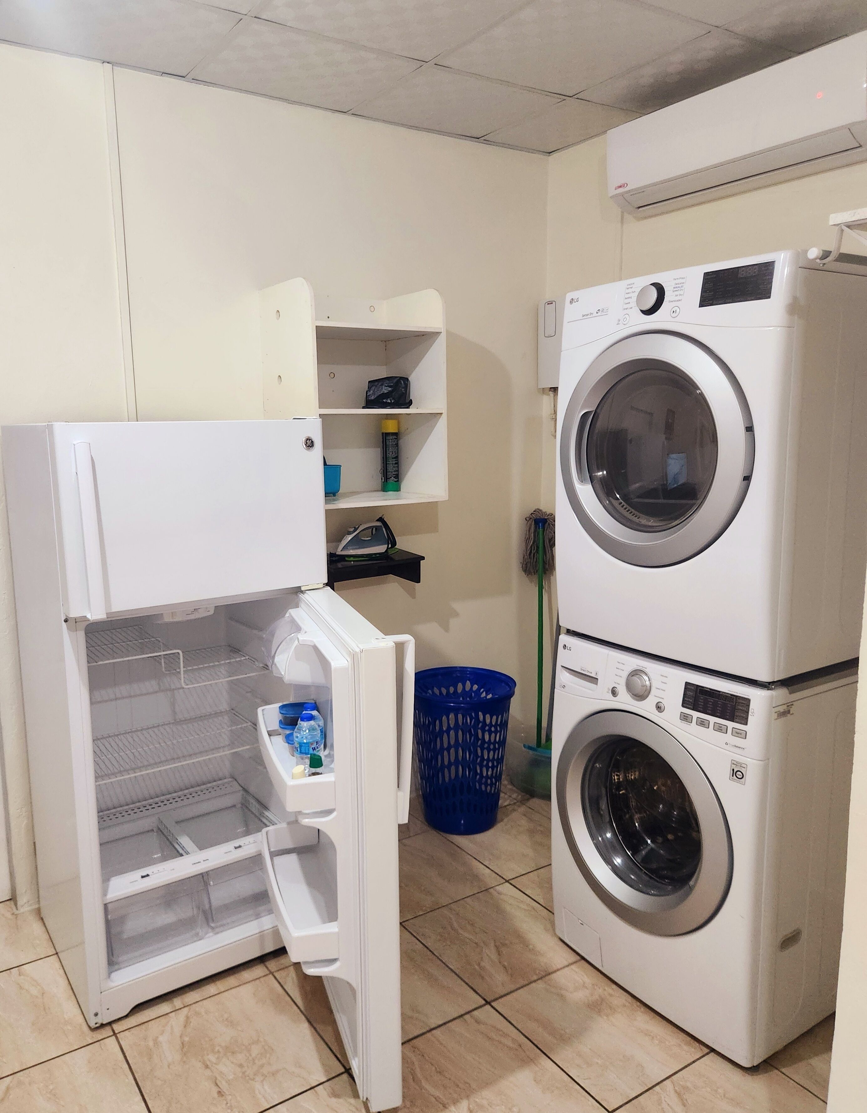 Un cuarto de lavandería con lavadora y secadora de carga frontal, un estante pequeño con una bolsa y una botella, y una cesta de ropa azul.