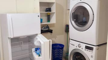 Un cuarto de lavandería con lavadora y secadora de carga frontal, un estante pequeño con una bolsa y una botella, y una cesta de ropa azul.