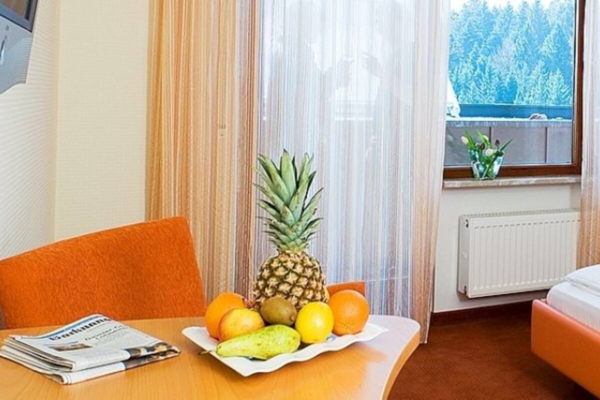 Quarto de hotel com cama, mesa com frutas e janela com vista para árvores.