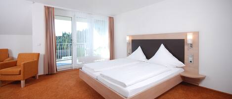 Ein Hotelzimmer mit Doppelbett, einem Sessel, einem Nachttisch und Blick ins Freie durch eine Schiebetür.